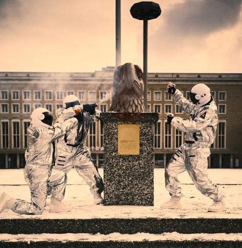 Die drei Hauptdarstellenden besprühen in Kosmonautanzügen den Kopf eines Reichsadlers.