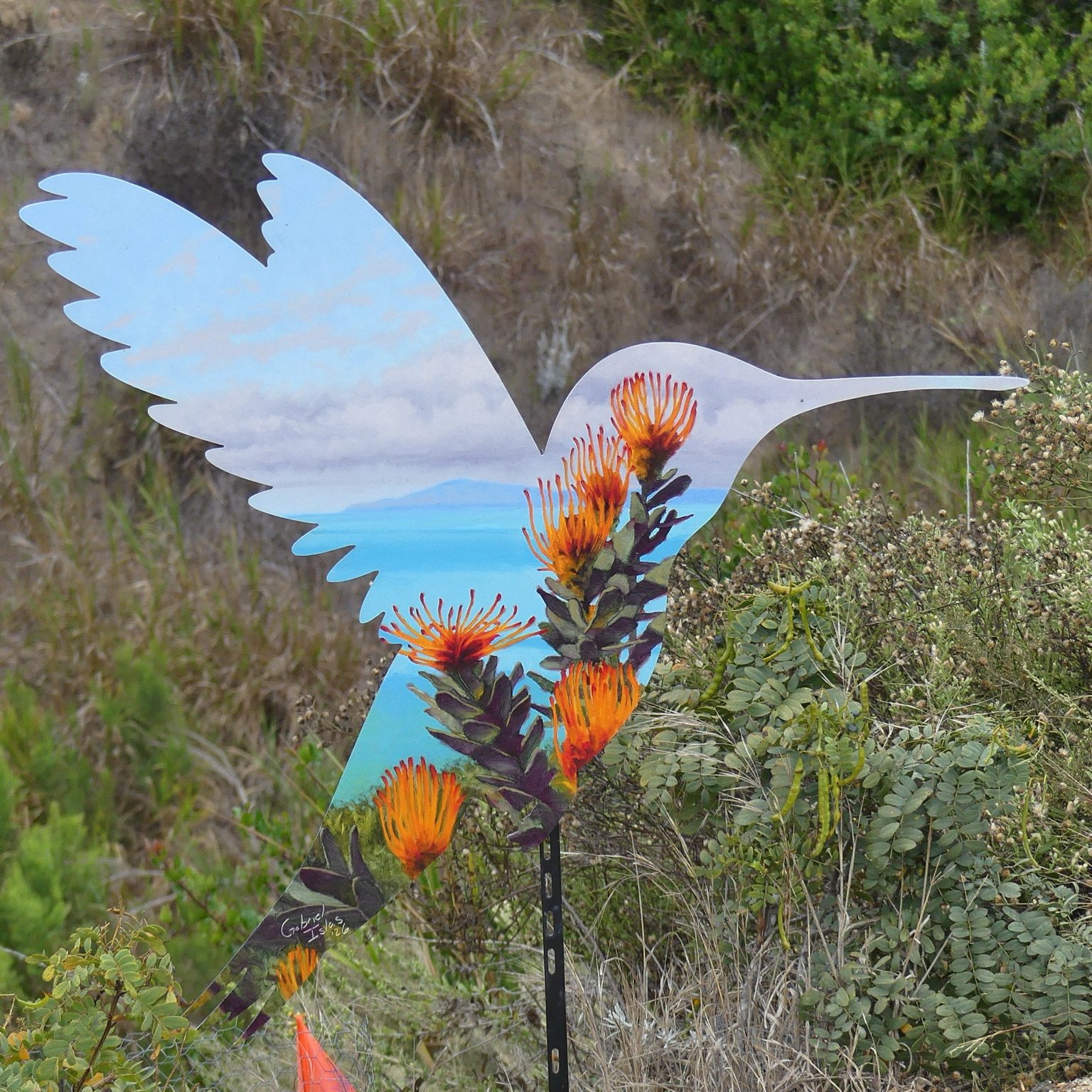 A hummingbird silhouette cutout painted with blue sky and orange cactus flowers against a backdrop of wild greenery