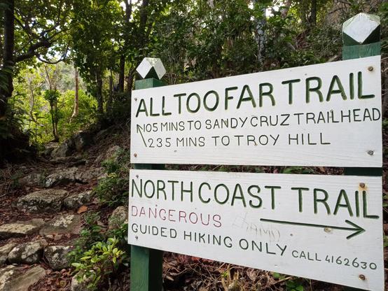 Big signs in the foreground: All too far Trail; North Coast Trail - Dangerous. hiking trail in the background.