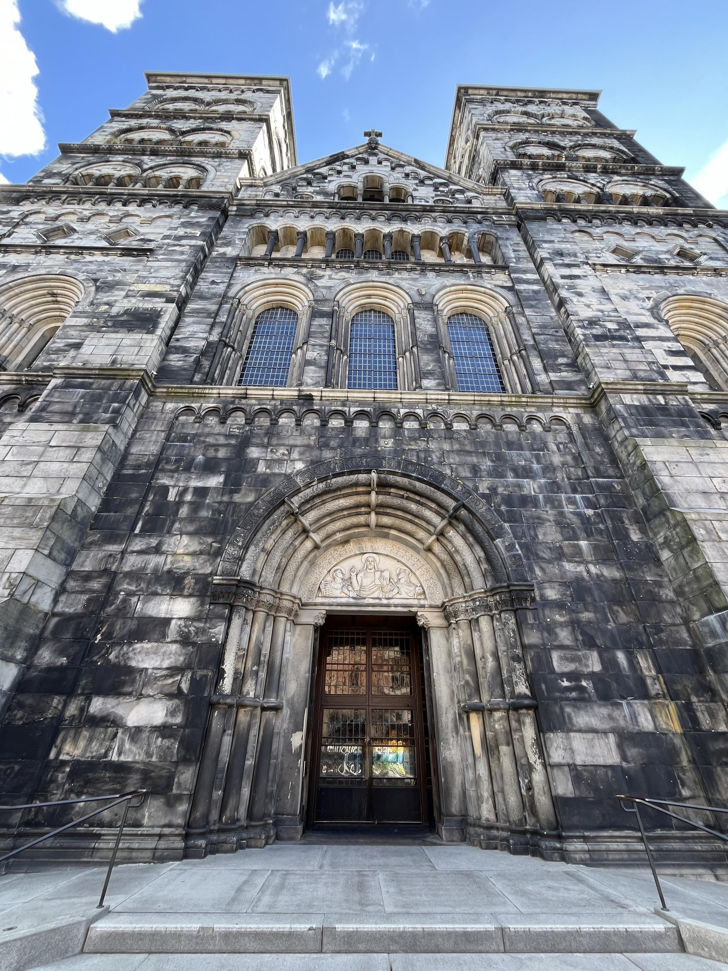 The image shows the entrance of a historic stone church with prominent architectural features, including two tall towers and decorative elements above the door. The sky is clear with a few clouds.